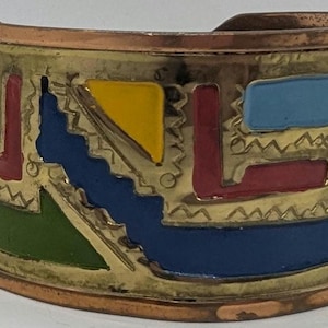 Op de afbeelding: Koperkleurige manchetarmband met een kleurrijk geometrisch ontwerp. De armband heeft vormen in rood, blauw, geel en groen, omlijnd in goud. Het ontwerp steekt af tegen een koperen achtergrond, wat een opvallend contrast creëert.