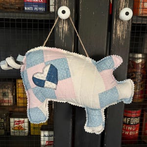 May include: A handmade, quilted pig-shaped wall hanging. The pig is made of patchwork fabric in shades of pink, blue, and white, with a heart-shaped patch. It is suspended by a natural twine string and has a ruffled edge.