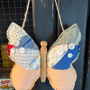 May include: A handmade butterfly decoration with patchwork wings and a wooden clothespin body. The wings are made of various patterned fabrics in blue, red, and green, with white buttons. The butterfly is suspended by a natural twine.