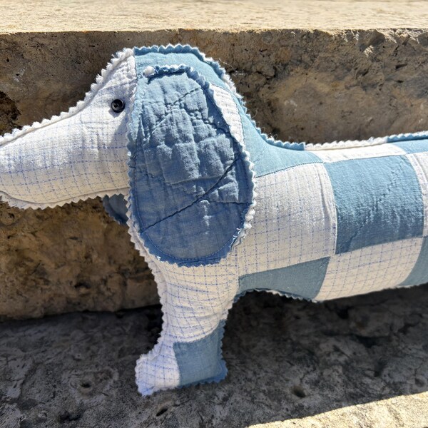 Könnte beinhalten: Ein Stoff-Dackel-Hundekissen mit Patchwork-Design. Der Hund hat ein blau-weiß kariertes Muster auf seinem Körper und seinen Ohren, mit einer weißen Schnauze und einem einzelnen schwarzen Auge. Das Kissen ist aus weichem Stoff gefertigt und hat ein handgefertigtes Aussehen.