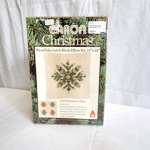 May include: A Caron Christmas Snowflake Latch Hook Pillow Kit. The kit includes a 14" x 14" (35.56 cm x 35.56 cm) design of a green snowflake on a cream background. The box features a pine needle border and the text "Latch Several for a Rug".