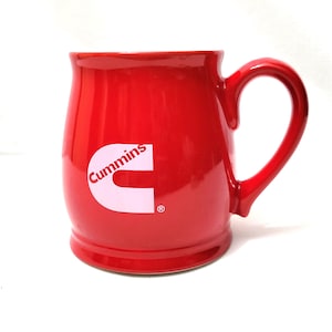 Könnte beinhalten: Eine glänzend rote Keramik-Tasse mit großem Henkel. Die Tasse zeigt das weiße "Cummins"-Logo auf der Vorderseite. Die Tasse hat eine abgerundete Form und einen breiten Boden.