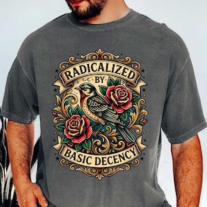 May include: Dark gray t-shirt featuring a detailed graphic. The design includes a bird, red roses, and the text "RADICALIZED BY BASIC DECENCY" in a banner. The artwork has a vintage tattoo style.