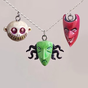 Puede incluir: Tres collares con colgantes con máscaras de personajes. Uno es una cara pálida sonriente con ojos morados y una sonrisa con dientes amarillos. Otro es una máscara verde con zarcillos negros. El tercero es una máscara de diablo roja con una sonrisa siniestra.