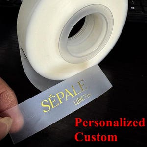 May include: A roll of clear tape with a transparent label. The label has gold text that reads "SÉPALE ЦВЕТЫ" and the words "Personalized Custom" in red. The tape is white and has a clear center.