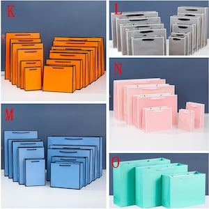 May include: Assortment of colorful paper gift bags in various sizes. The bags are made of sturdy paper and have black handles. The bags are arranged in rows, with the largest bags at the back and the smallest bags at the front. The bags are available in orange, blue, pink, gray, and green.