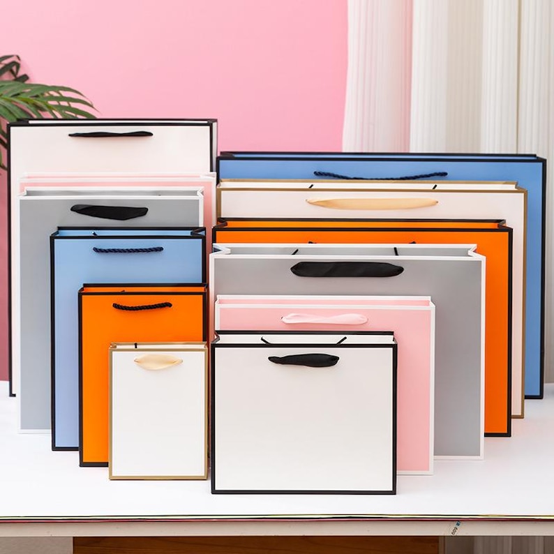 May include: A set of colorful paper gift bags with black trim and handles. The bags are arranged in a stack, with the largest bag at the bottom and the smallest bag at the top. The bags are all different colors, including blue, orange, pink, and gray.