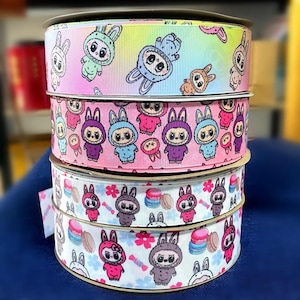 Könnte beinhalten: Vier Rollen dekoratives Band mit sich wiederholenden Cartoon-Hasenfiguren. Die obere Rolle hat einen Regenbogen-Farbverlauf, die zweite ist rosa und die unteren beiden sind weiß. Die Bänder zeigen Hasen in verschiedenen Farben und Outfits.