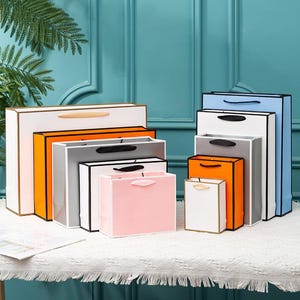 May include: A set of six colorful paper gift bags in various sizes. The bags are made of sturdy paper and have black handles. The bags are arranged in a stack, with the largest bag at the bottom and the smallest bag at the top.