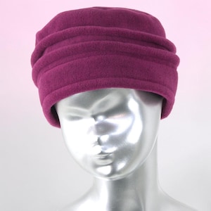 Peut inclure: Un bonnet en polaire magenta profond doté d'une calotte arrondie et de trois bandes de tissu froncées horizontales entourant la base. La matière présente une texture douce et duveteuse, offrant une coupe structurée autour de la tête.
