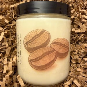 May include: A glass jar candle with a black lid. The label shows three brown coffee beans. The text "Coffee Bean luminaire candle co." is printed vertically on the left. The candle is on a bed of brown paper.