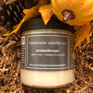 May include: A white soy candle in a glass jar with a black lid. The label reads "luminaire candle co. aromatherapy gluten lead phthalate free soy candle etsy.com/shop/luminairecandleco".