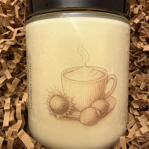 May include: A cylindrical glass jar candle with a black lid. The label features a sepia-toned illustration of a coffee cup, hazelnuts, and the words "Hazelnut Cappuccino". The candle sits in brown packing material.