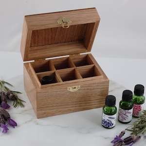 May include: A wooden essential oil storage box with six compartments, open to reveal the interior. The box is made of light-coloured wood and has a brass clasp. Three green essential oil bottles are next to the box.