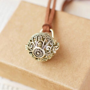 May include: A gold-toned, spherical pendant necklace with an ornate filigree design. The pendant hangs from a brown leather cord. The intricate metalwork features floral and swirling patterns. The necklace is displayed against a neutral background.