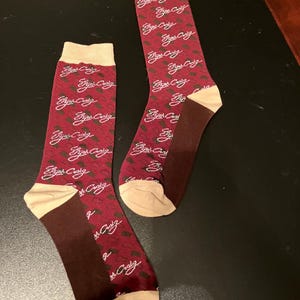 May include: A pair of maroon socks with a white "Elijah Craig" logo repeated all over the sock. The socks have a tan colored toe and heel.