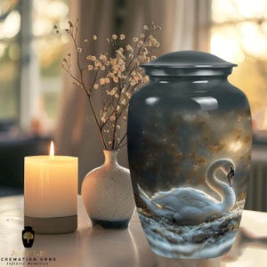 Puede incluir: Una urna funeraria gris oscuro con una ilustración de un cisne. La urna tiene tapa y es cilíndrica. El cisne es blanco con cuello largo y descansa sobre un nido. Una vela encendida y un pequeño jarrón con flores están junto a la urna. El texto "APPLE CREMATION URNS Infinite Memories" está en la parte inferior.