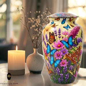 May include: A decorative cremation urn with a yellow background, adorned with colorful butterflies and flowers. The urn features a lid and is surrounded by a lit candle, a small vase with flowers, and a small vase.