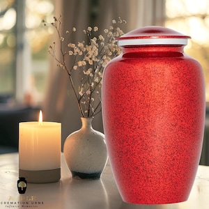May include: Red metal cremation urn with a silver lid. The urn has a speckled finish and is sitting on a table with a candle and a vase of flowers.