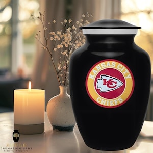 Op de afbeelding: Zwarte crematieurn met een witte rand en het Kansas City Chiefs-logo. Naast de urn staan een brandende kaars en een kleine vaas met bloemen. De urn heeft de tekst "AIPLE CREMATION URNS INFINITE MEMORIES" aan de onderkant.