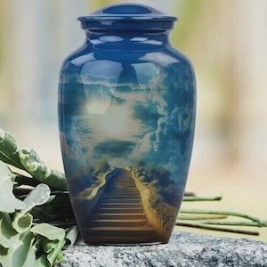 May include: A blue cremation urn with a lid, featuring a scenic painted design of a pathway leading to the sky. The urn is glossy and sits on a stone surface, with greenery visible on the side.