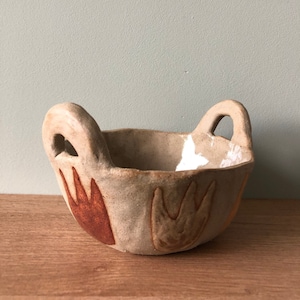 Könnte beinhalten: Eine kleine, beige Keramik Schüssel mit zwei Griffen. Die Schüssel hat eine strukturierte Oberfläche und ist mit braunen und orangefarbenen abstrakten Mustern verziert.