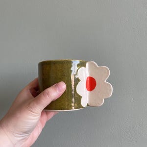 Puede incluir: Una taza de cerámica verde con un asa de flor blanca. La flor tiene un centro rojo.