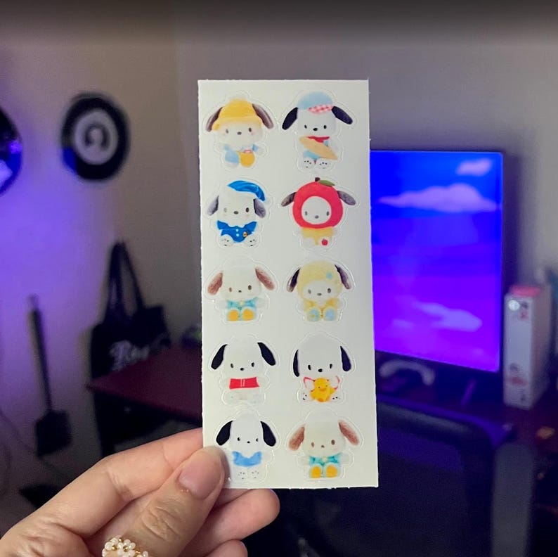 May include: A sheet of stickers featuring cartoon dog characters in various outfits. The stickers are white with colorful illustrations of the dogs wearing hats, holding objects, and dressed in different outfits. The stickers are held by a person's hand.
