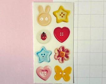 May include: A rectangular sticker sheet featuring eight individual button-shaped designs. The stickers include a beige rabbit, a yellow star, a white circle with a ladybug, a red heart, a light blue star with a face, a pink flower, a pink heart, and a yellow butterfly.