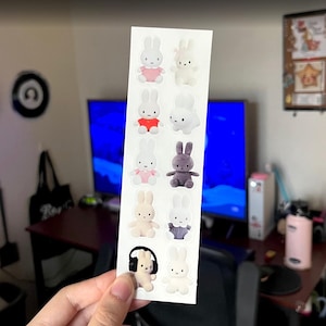 May include: A vertical sticker sheet featuring ten individual die-cut stickers of Miffy the rabbit in various outfits and poses, including a white rabbit wearing pink, red, or blue clothing, a striped grey rabbit, and a rabbit wearing black headphones.