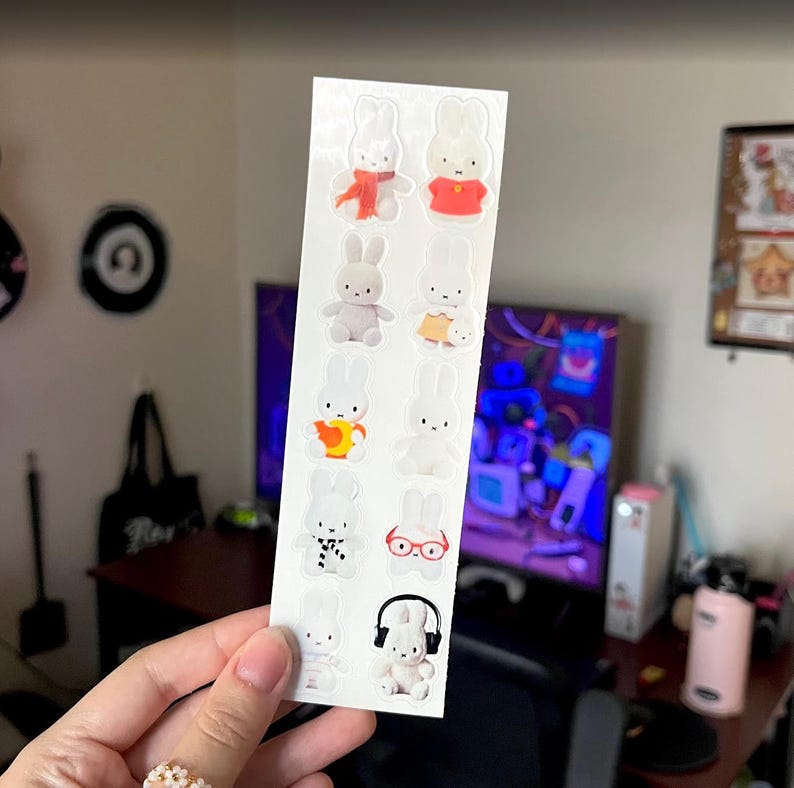 May include: A vertical sticker sheet featuring ten individual white rabbit characters in various outfits and poses. The rabbits are depicted wearing items such as a red scarf, a red shirt, a yellow skirt, glasses, and black headphones, all on a white background.