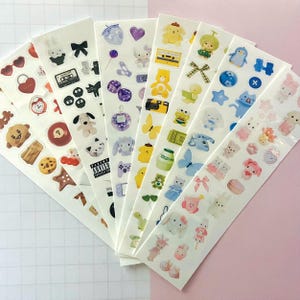 May include: A set of ten sheets of colorful stickers featuring various cute cartoon characters, animals, and objects. The stickers are on a white background and include designs like a bear, a penguin, a butterfly, a camera, a milk carton, a lollipop, a cassette tape, a video game controller, and a heart.