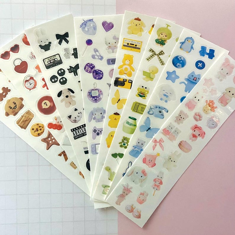 May include: A set of ten sheets of colorful stickers featuring various cute cartoon characters, animals, and objects. The stickers are on a white background and include designs like a bear, a penguin, a butterfly, a camera, a milk carton, a lollipop, a cassette tape, a video game controller, and a heart.