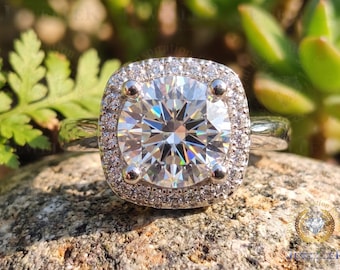 May include: A close-up of a diamond ring with a square halo setting. The large, round-cut diamond is surrounded by smaller stones, set on a silver band. The ring is displayed on a textured stone surface, with greenery in the background.