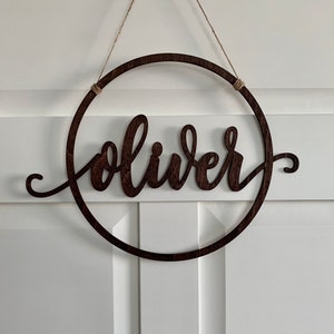 Op de afbeelding: Een houten cirkel met de naam "Oliver" uitgesneden in een cursief schrift. De cirkel hangt aan een touwtje.