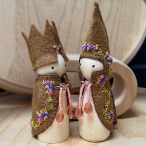 May include: Two wooden peg dolls, one with a crown and the other with a pointed hat, both wearing brown capes with floral embroidery. The dolls have white faces and are adorned with pink beaded accents, creating a whimsical, handcrafted aesthetic.