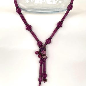 Könnte beinhalten: Eine violette Perlenkette mit einem Perlencluster in der Mitte und einem langen Quastenanhänger. Die Kette wird auf einer durchsichtigen Glasvase präsentiert.