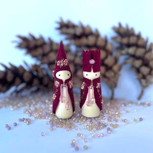 May include: Two handcrafted wooden dolls. They have burgundy felt capes and hats, decorated with floral embroidery. The dolls have white faces with simple features and pink beaded accents. The background includes pinecones and scattered crystals.