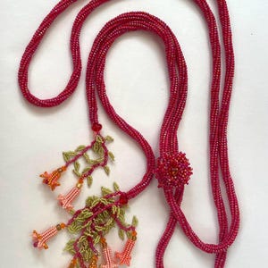 Könnte beinhalten: Eine lange Perlenkette in Pink- und Rottönen. Die Kette hat einen floralen Anhänger mit rosa, orangefarbenen und grünen Perlen.