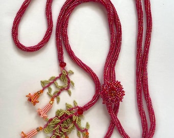 May include: A long, beaded necklace with a pink and red color scheme. The necklace has a floral pendant with pink, orange, and green beads.