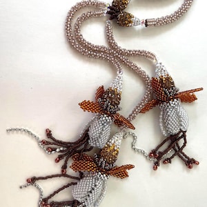Könnte beinhalten: Eine Halskette mit Perlen und einem floralen Design. Die Halskette besteht aus Strängen kleiner, hellbrauner Perlen. Der Mittelpunkt umfasst silberne, braune und goldfarbene, perlenbesetzte blütenartige Formen mit hängenden Akzenten. Die Halskette wird auf einer weißen Oberfläche präsentiert.