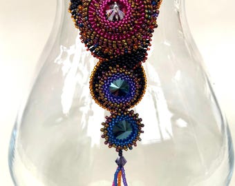 May include: A beaded necklace with a multi-coloured, circular pendant and a tassel. The pendant features a central crystal and is surrounded by smaller beads in shades of pink, orange, and gold. The tassel is made of colourful beads and hangs from the bottom of the pendant.