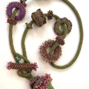 Puede incluir: Un collar de cuentas verdes con múltiples colgantes de cuentas con forma de flor. El collar está hecho con pequeñas cuentas verdes y tiene acentos rosas y morados.