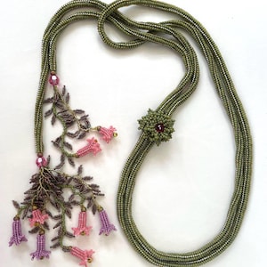 Könnte beinhalten: Eine grüne Perlenkette mit einem Blumenanhänger und einem Perlenquaste mit rosa und violetten Blüten.