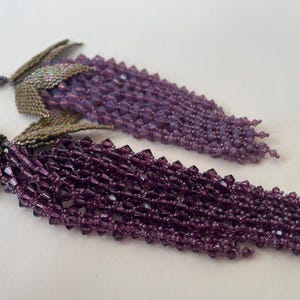 Könnte beinhalten: Ein Paar Perlenohrringe in einem violetten und grünen Farbschema. Die Ohrringe bestehen aus kleinen Perlen, die miteinander verwoben sind, um einen fransigen Effekt zu erzielen. Die Ohrringe sind ein einzigartiges und stilvolles Accessoire.