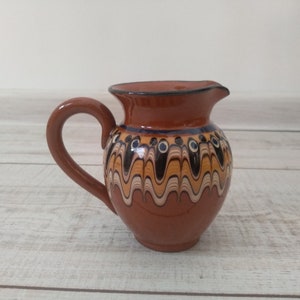 Può includere: Un piccolo brocca in ceramica marrone con un manico. La brocca ha un design decorativo di linee ondulate e cerchi in tonalità di marrone, blu e bianco.
