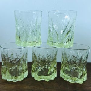 May include: Five clear drinking glasses with a textured, bark-like design. The base of each glass has a pale yellow-green tint. The glasses are arranged in a pyramid shape on a dark wooden surface.