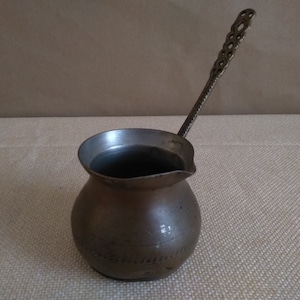 May include: A vintage, silver-toned, Turkish coffee pot with a long, ornate handle. The pot has a rounded body and a small spout.