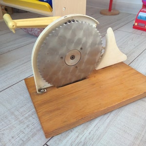 Könnte beinhalten: Ein Vintage-Spielzeug-Brotschneider mit cremefarbenem Rahmen, einer großen Kreissäge und einer Holzbasis. Der Griff und der Klingenschutz sind ebenfalls cremefarben. Das Spielzeug soll das Schneiden von Brot simulieren.