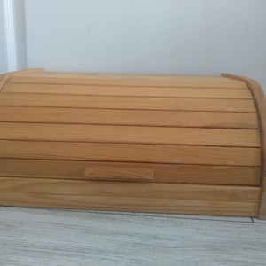 Può includere: Un porta pane in legno con un coperchio incernierato e un design semplice. Il coperchio ha un design a doghe e la scatola è in legno naturale.